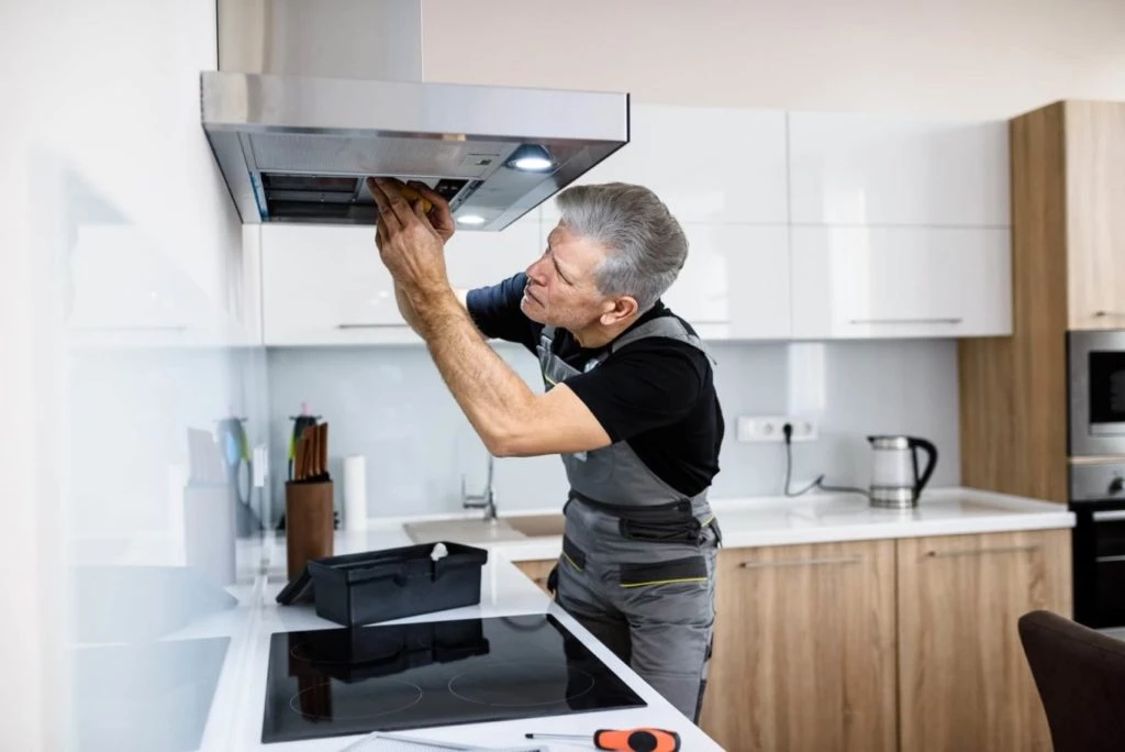 Aged repairman in uniform working, fixing broken kitchen extractor fan using screwdriver. Commercial hood repair service concept.